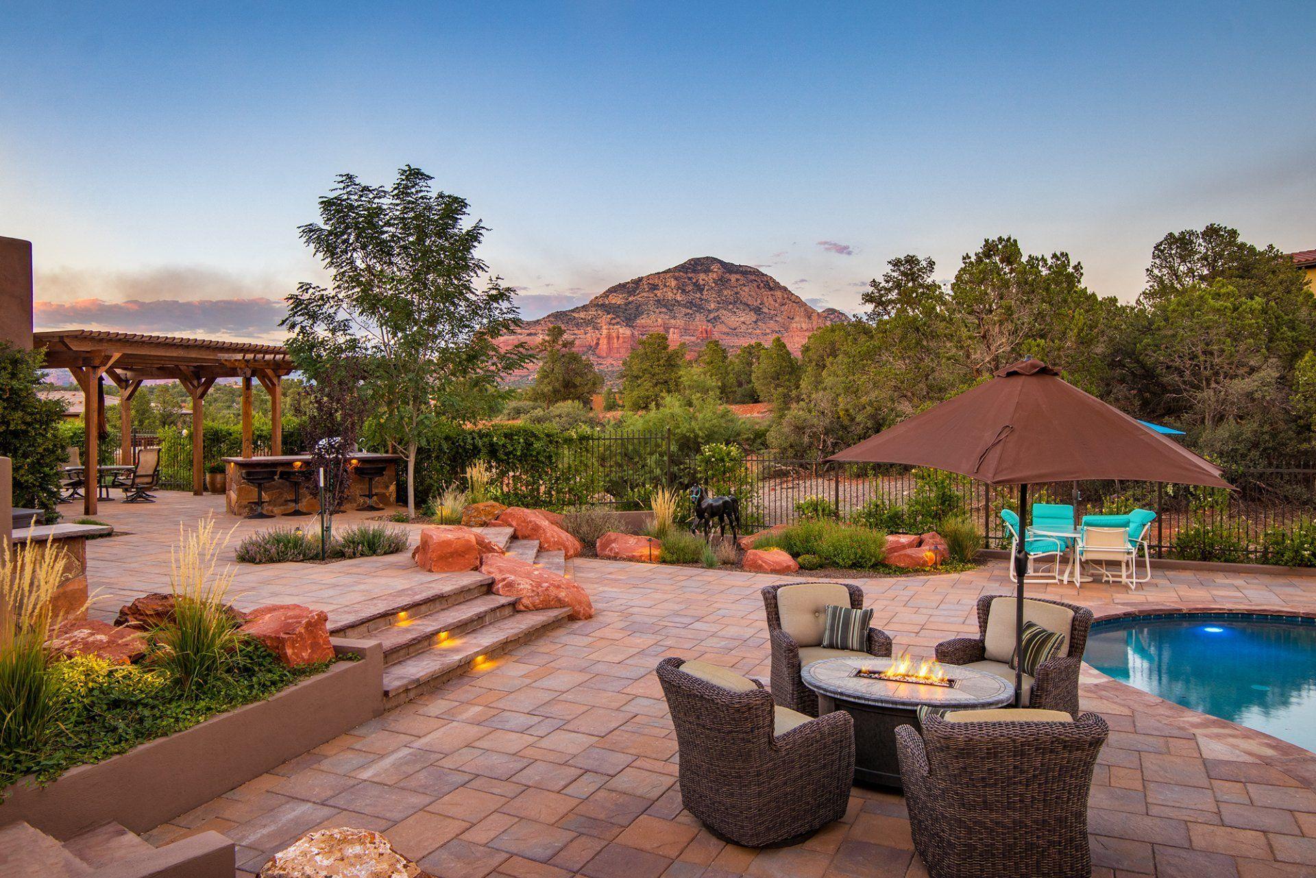 A patio with a table , chairs , umbrella and a pool with a mountain in the background.