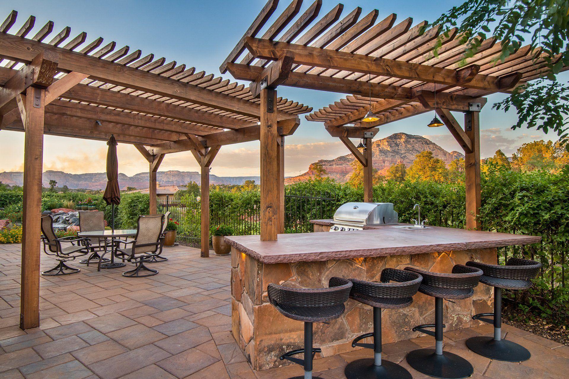 Pergola-covered stone bar with stools and a grill on a paver patio at twilight