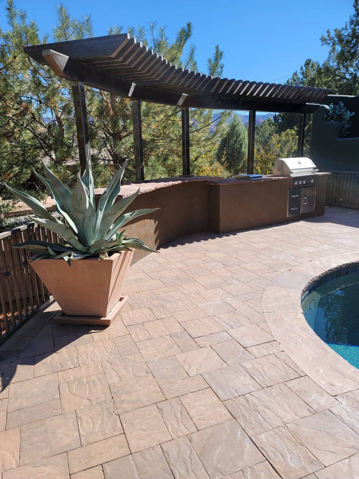 Outdoor kitchen with built-in grill under a pergola beside a pool and potted agave