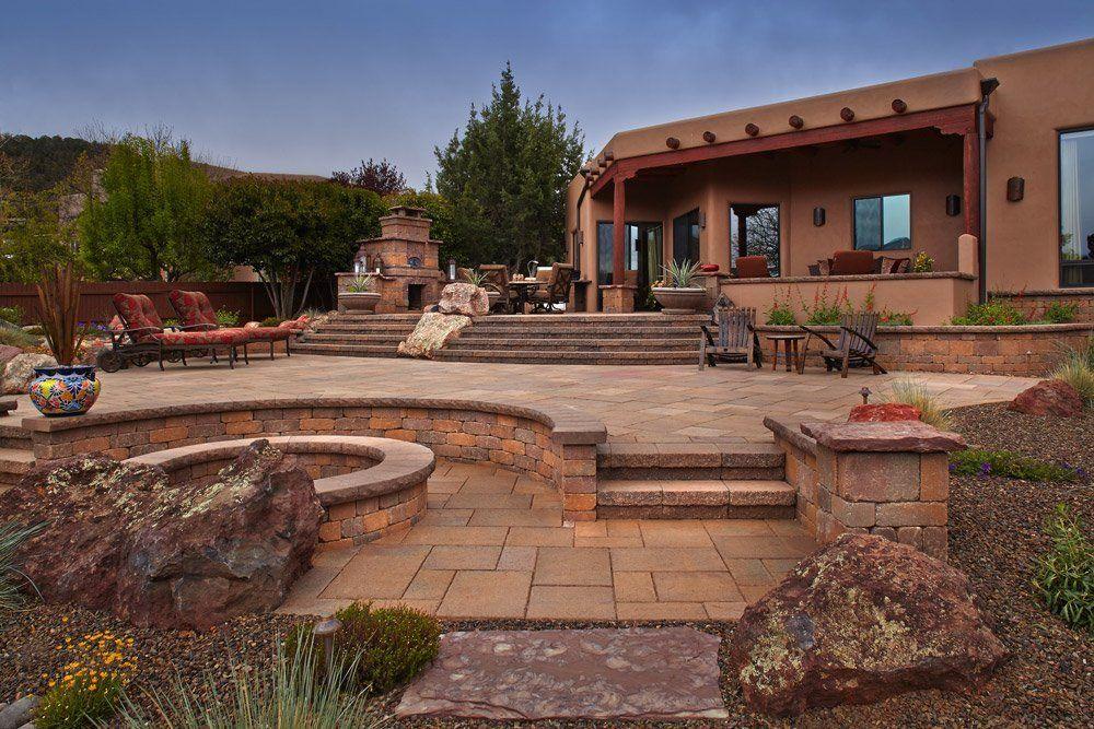 Paved patio with built-in fire pit, steps, and southwestern-style home. Landscaping and lounge chairs are present.