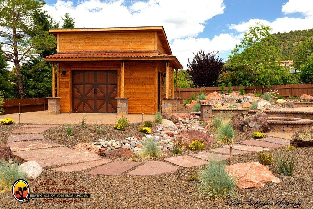 A wooden garage with a brown garage door is surrounded by rocks and trees.