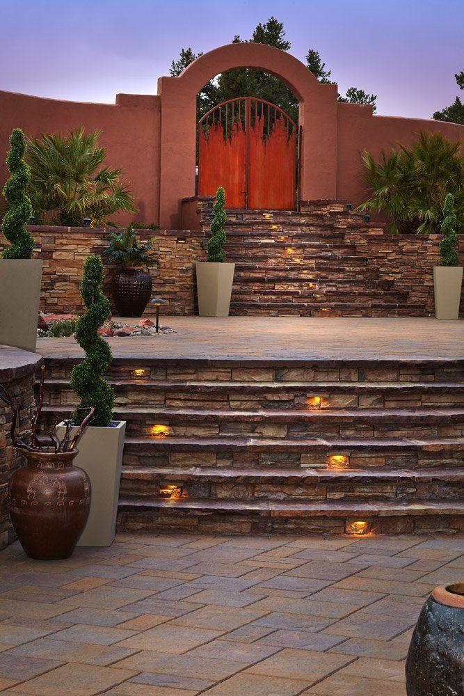 Stone steps lead to a courtyard with a rust-colored gate under an arch; potted plants and glowing lights.